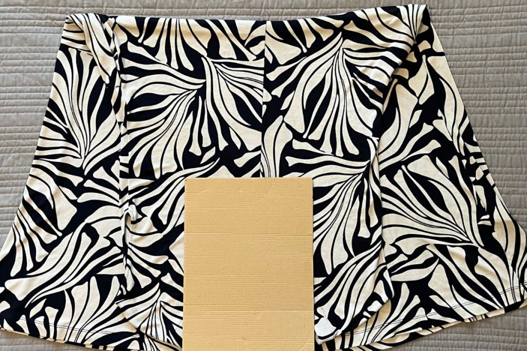 A black and white patterned dress is laid out, folded in half with a smaller rectangular piece of brown cardboard on top at the neckline.