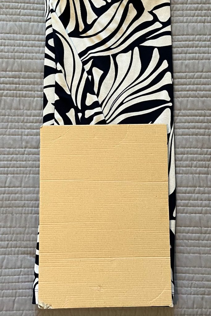 A black and white patterned dress is laid out, folded in thirds vertically with a smaller rectangular piece of brown cardboard on top at the fold. Some of the dress is still visible at the top.