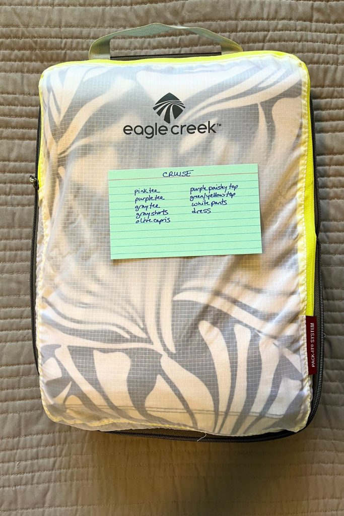 A closed, white packing cube is filled with clothes, with a black and white patterned item seen at the top. The words Eagle Creek and a logo are in black at the top of the cube. A green index card with handwritten list of items in the cube is lying on top of the closed packing cube.