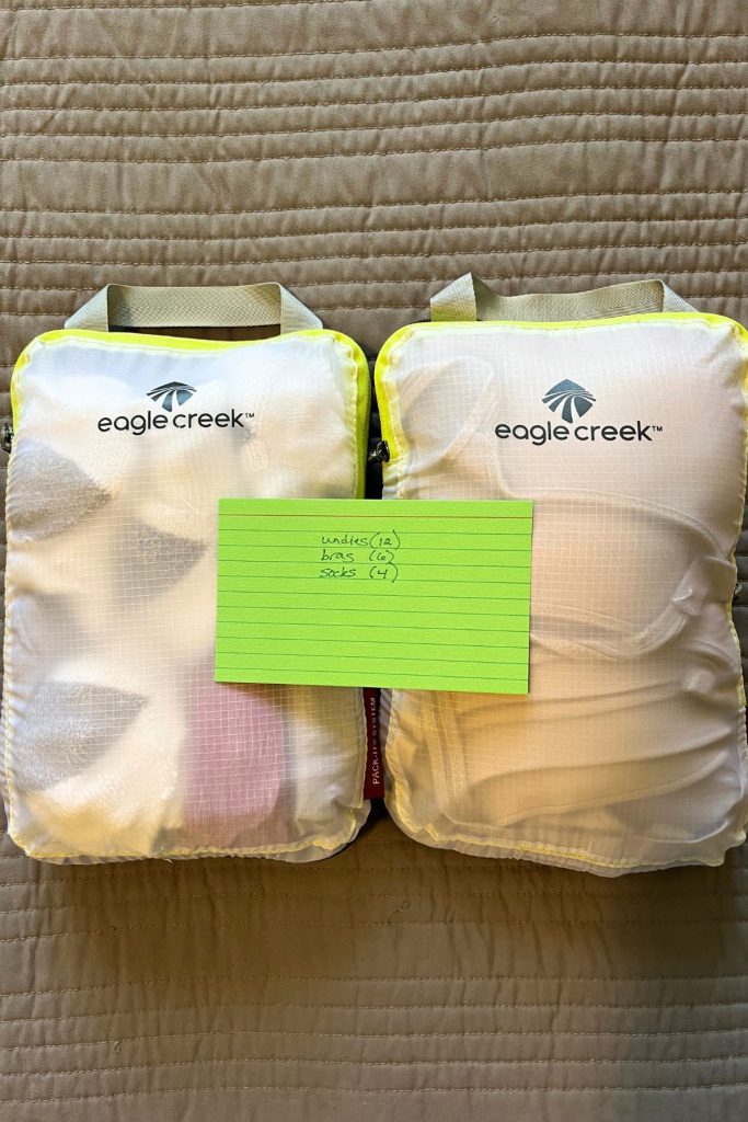 Two closed, white packing cube are filled with clothes, with a black and white patterned item seen at the top. The words Eagle Creek and a logo are in black at the top of each cube. A green index card with handwritten list of items in the cube is lying across the closed packing cubes.