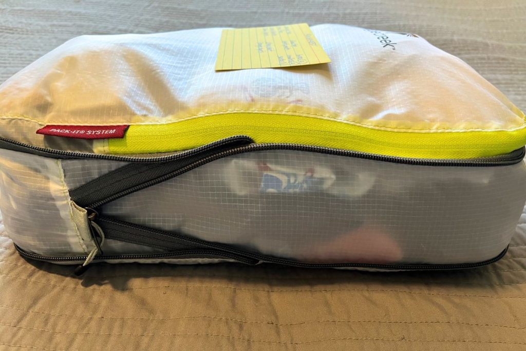 A closed, white packing cube is filled with clothes, and is viewed from the side. A yellow index card with a handwritten list of items in the cube is lying on top of the closed packing cube.