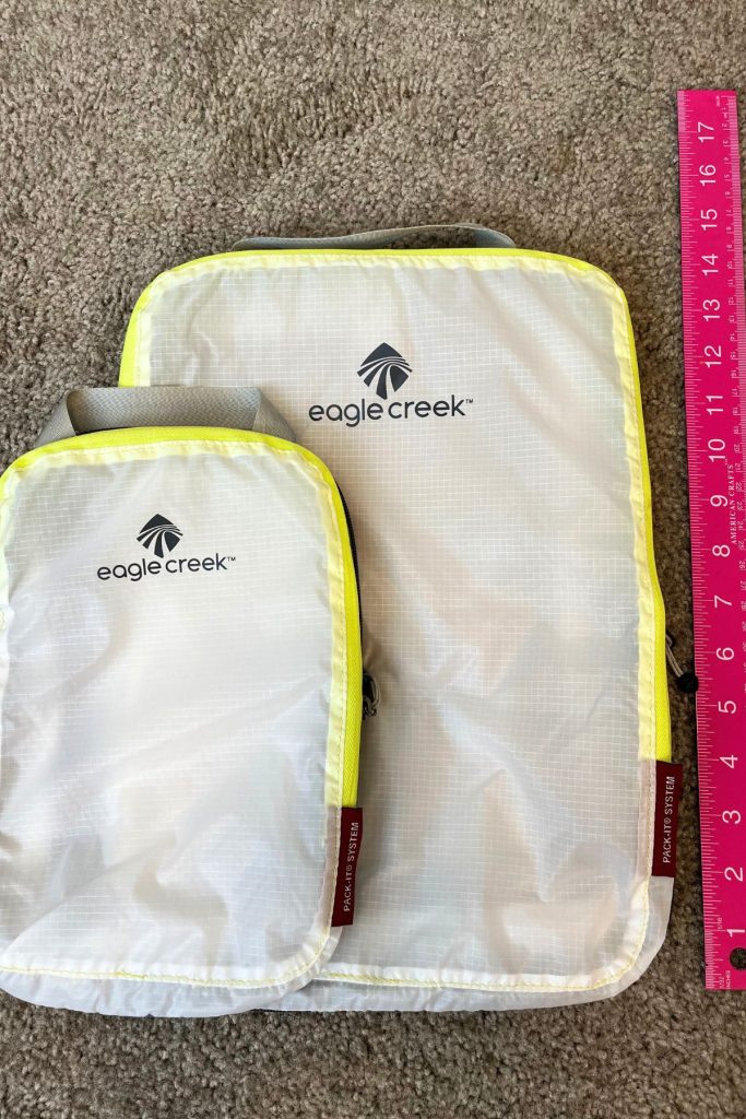 Two white packing cubes, a large one with a smaller one lying on top. Each cube has the words Eagle Creek and a logo in black. A pink ruler is on the right, showing that the larger cube is 14 inches long.