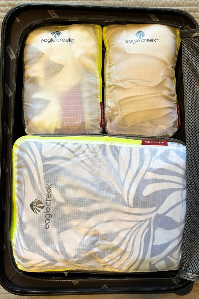 Three white packing cubes, two small ones at the top and one large one at the bottom, are placed inside the open half of a suitcase. Each packing cube has the words Eagle Creek and a logo on it in black.