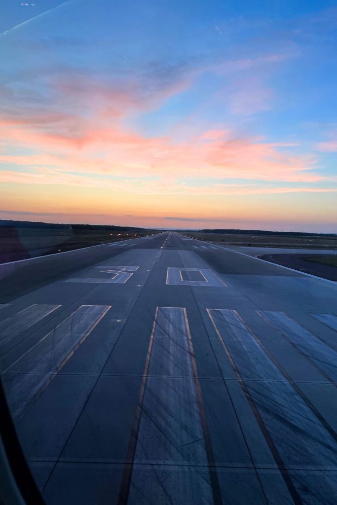 Looking down an airport runway from the window of an airplane, while the sun is setting in the background. The runway has a large number 30 painted on it in white, along with several large vertical stripes at the bottom.