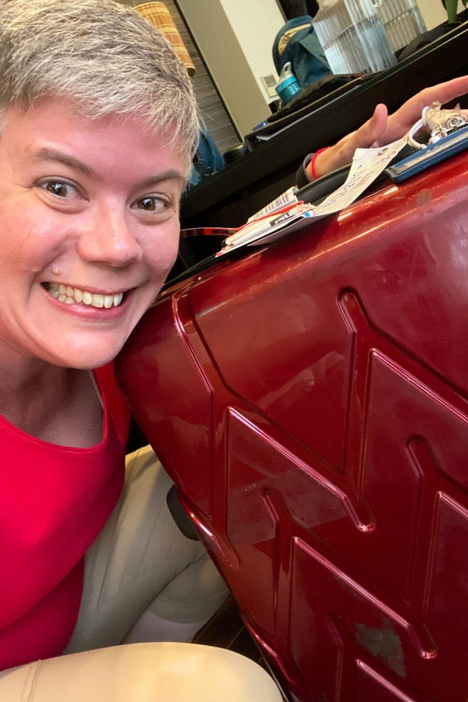 A smiling woman, wearing a red shirt and tan pants, crouches next to a large maroon-colored suitcase. There are several paper airline tags attached to the suitcase handle.