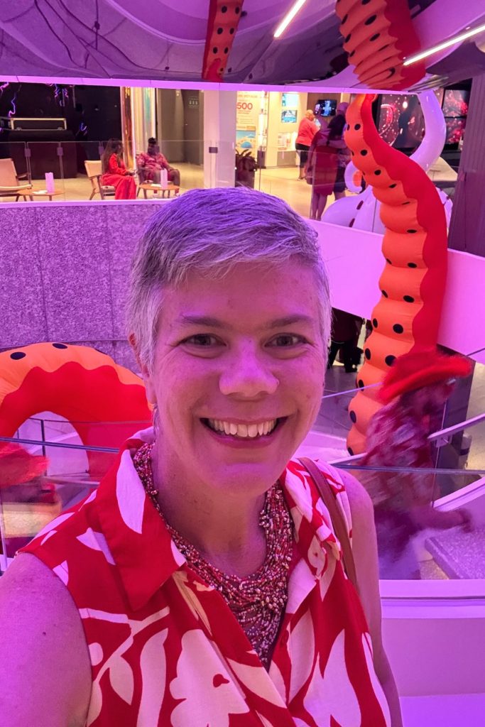 A smiling woman, wearing a red and white print sleeveless top and a red beaded necklace stands in front of an atrium on a Virgin Voyages cruise ship. The atrium below her has two large, red inflatable octopus tentacles.