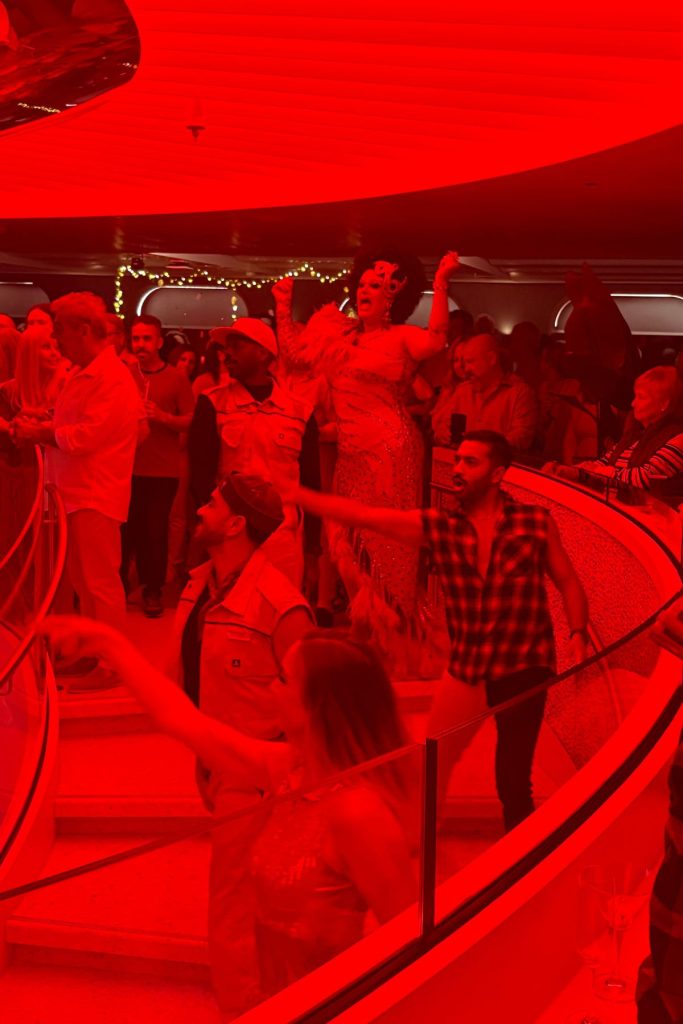 Several people are standing on a large spiral staircase, including a drag queen, three male performers, and one female performers. They are all singing and dancing. The entire room is bathed in red light.