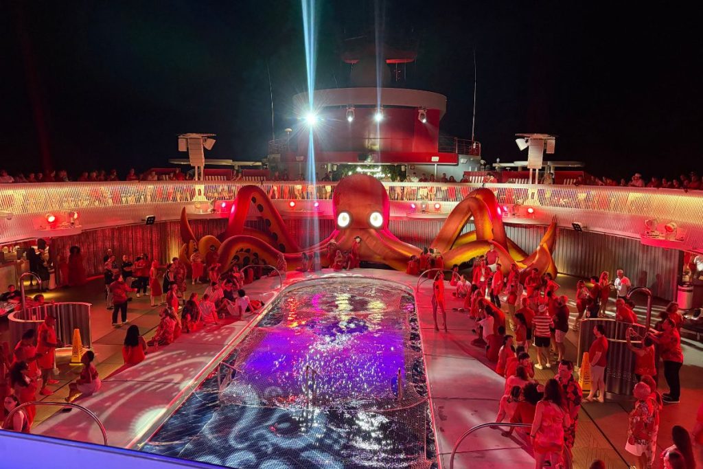 The pool deck on a Virgin Voyages cruise on Scarlet Night. A large, red inflatable octopus sits on one end of the pool, its eyes lit up with white lights. There is a crowd of people around the pool itself, along with more people on the raised deck area above the pool.