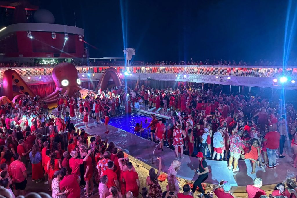 The pool deck on a Virgin Voyages cruise on Scarlet Night. A large, red inflatable octopus sits on one end of the pool, its eyes lit up with white lights. There is a crowd of people in and around the pool itself, along with more people on the raised deck area above the pool.