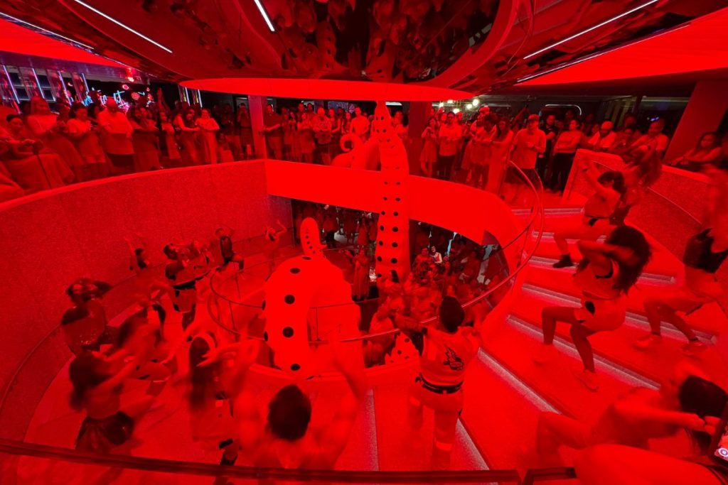 A large group of performers are on different steps of a large spiral staircase. Each performer is gesturing wildly mid-dance. A large audience surrounds the top of the staircase, watching the performers. The entire scene is bathed in red light.