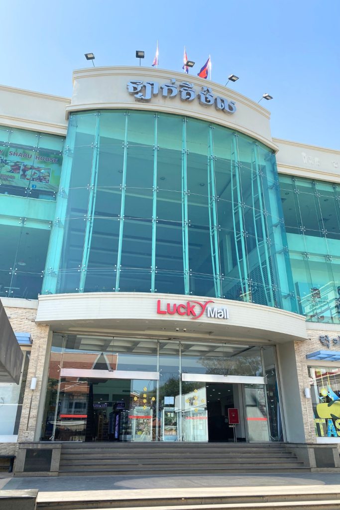 Glass-fronted shopping mall with a red and white Lucky Mall sign above the entrance, Khmer lettering and three flags on the roof, and a restaurant banner on the left, under a clear blue sky.