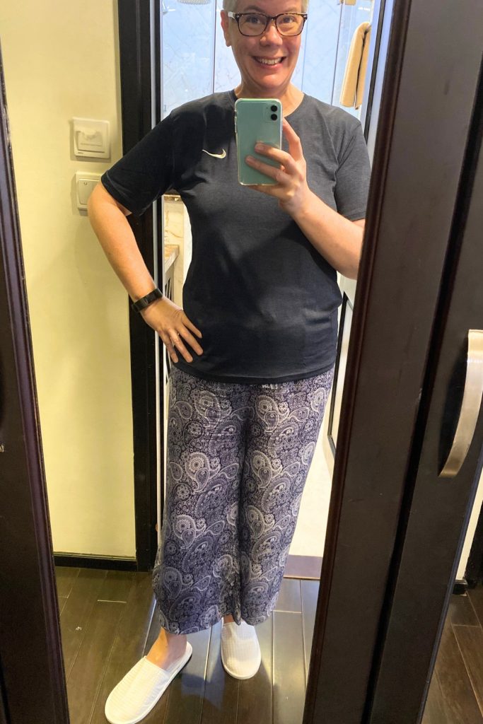 Woman with short gray hair and glasses taking a mirror selfie in a hotel room, wearing a navy T-shirt, blue and white paisley pants, and white hotel slippers.