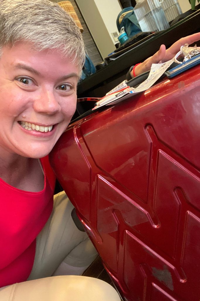 Smiling woman in a red top leaning her head against a glossy red hard-shell suitcase with luggage tags and a blue name tag on top.