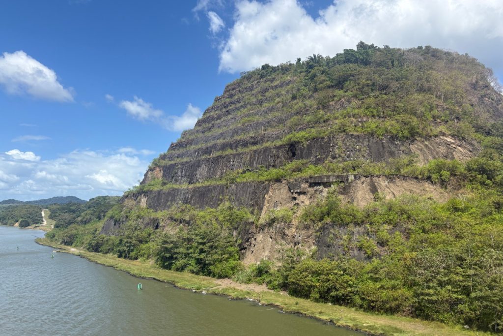 Steep terraced hillside of bare rock layers and green brush beside a calm channel under a blue sky