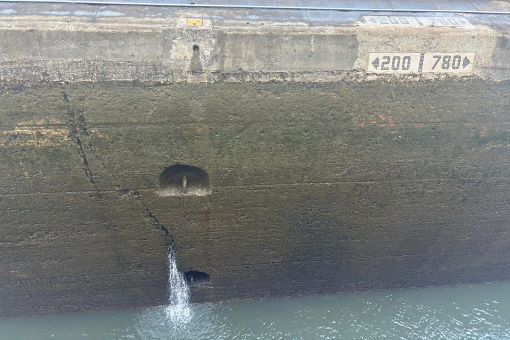 Weathered concrete lock wall with a crack and stains, a small stream of water running out near the waterline, and distance markers reading 200 and 780 along the top