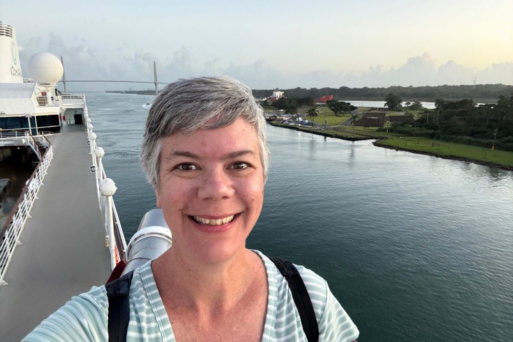Woman taking a selfie on a ship's top deck at sunrise, with a cable-stayed bridge and green shoreline behind her