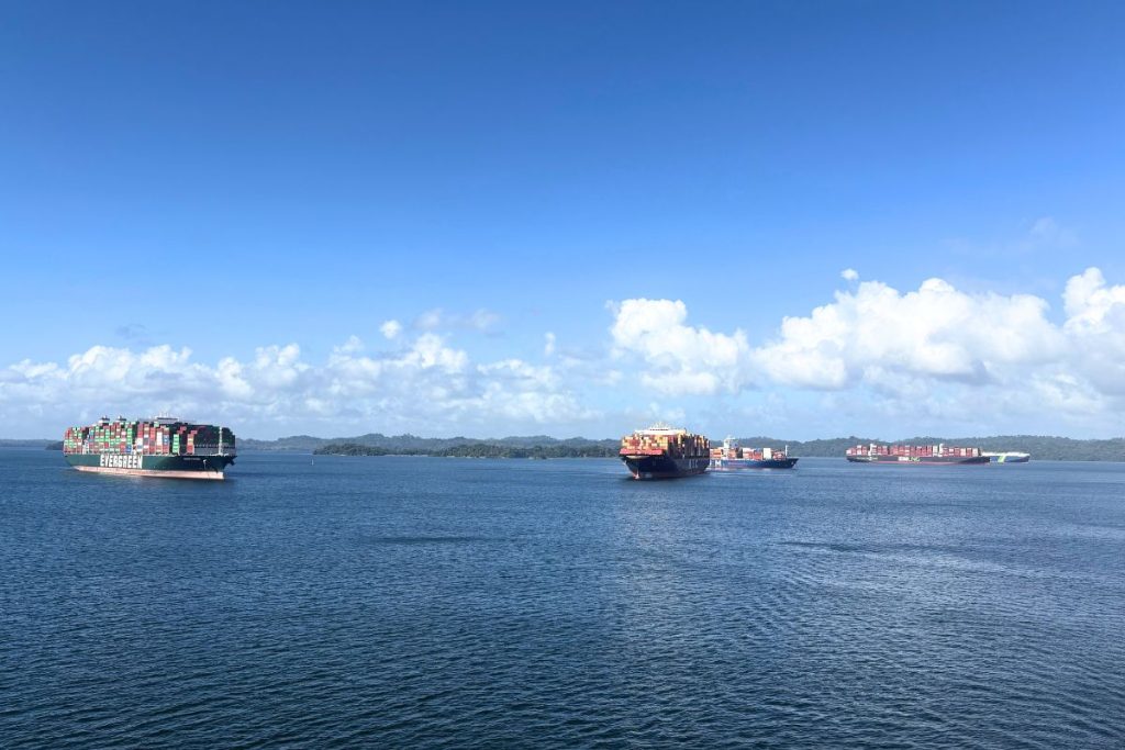 Four container ships loaded with stacked containers on calm blue water under a cloudy blue sky