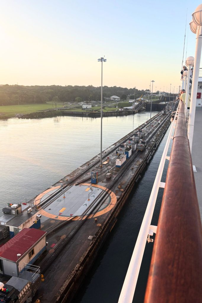 View from a ship's railing down onto a lock wall with tracks, several small electric locomotives, and a circular orange platform