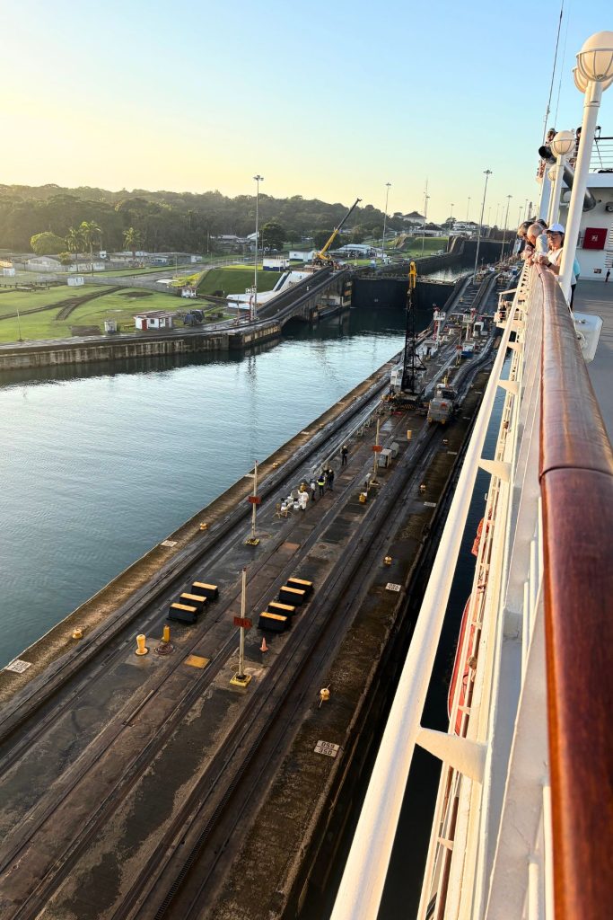 View from a ship's railing down a lock wall with tracks, numbered posts, a crane, and workers in safety gear
