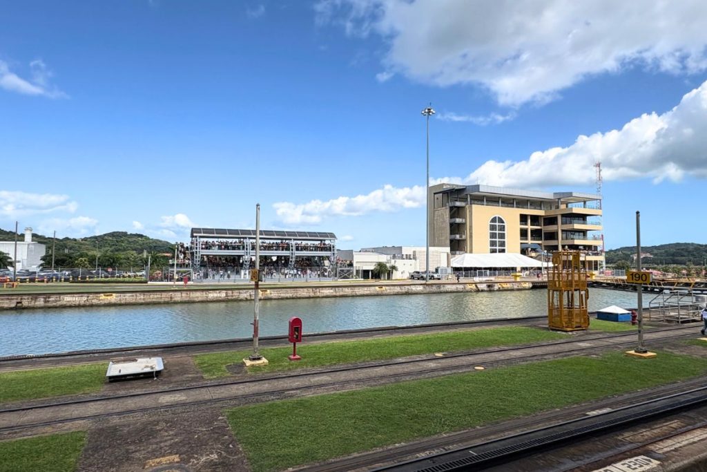 View across a lock wall to a multi-level visitor center and a crowded observation deck under a blue sky