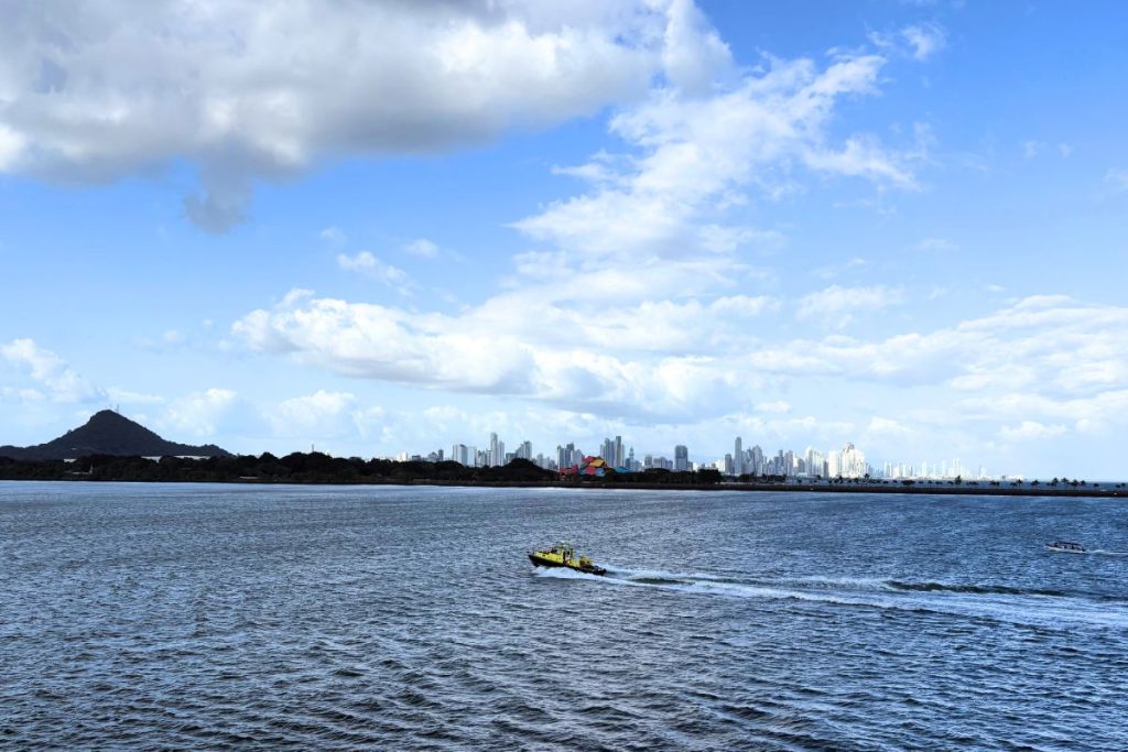 Panama City skyline across blue water, with a small yellow boat crossing in front and a hill at left