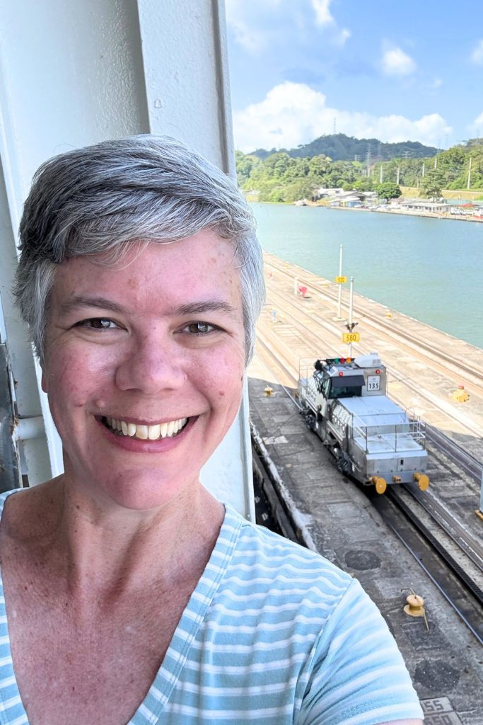 Smiling woman taking a selfie on a ship, with an electric locomotive numbered 135 on the lock wall below