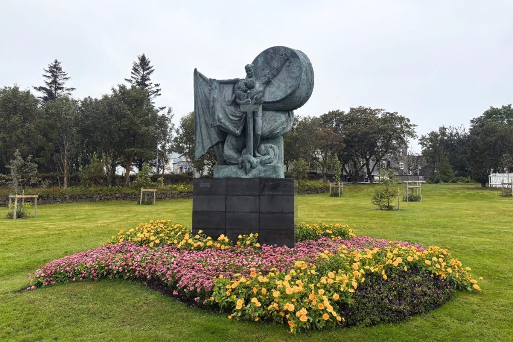 Green-patina statue on a dark pedestal ringed with pink and yellow flowers near Tjörnin lake