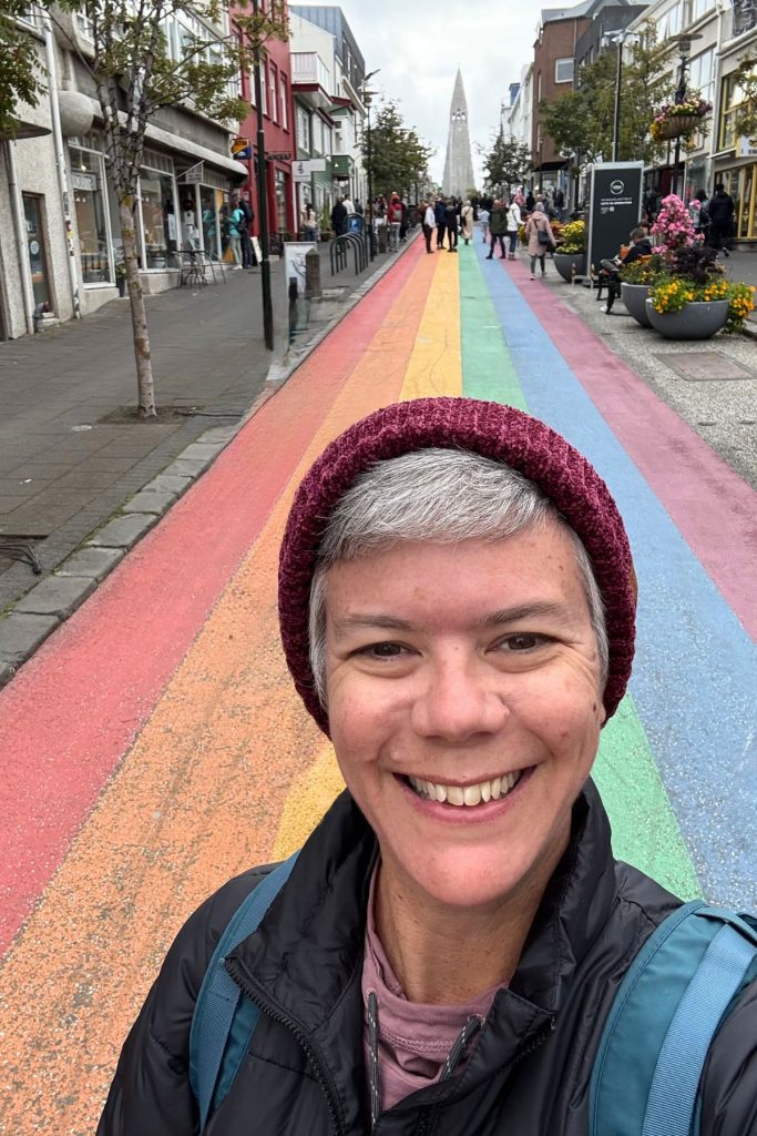 Woman in a burgundy hat on a rainbow-painted street with Hallgrímskirkja in the distance