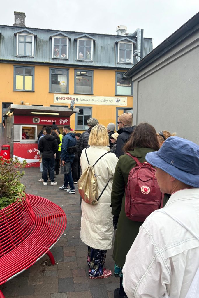 Line of people in jackets waiting at a small red hot dog stand beside a red bench