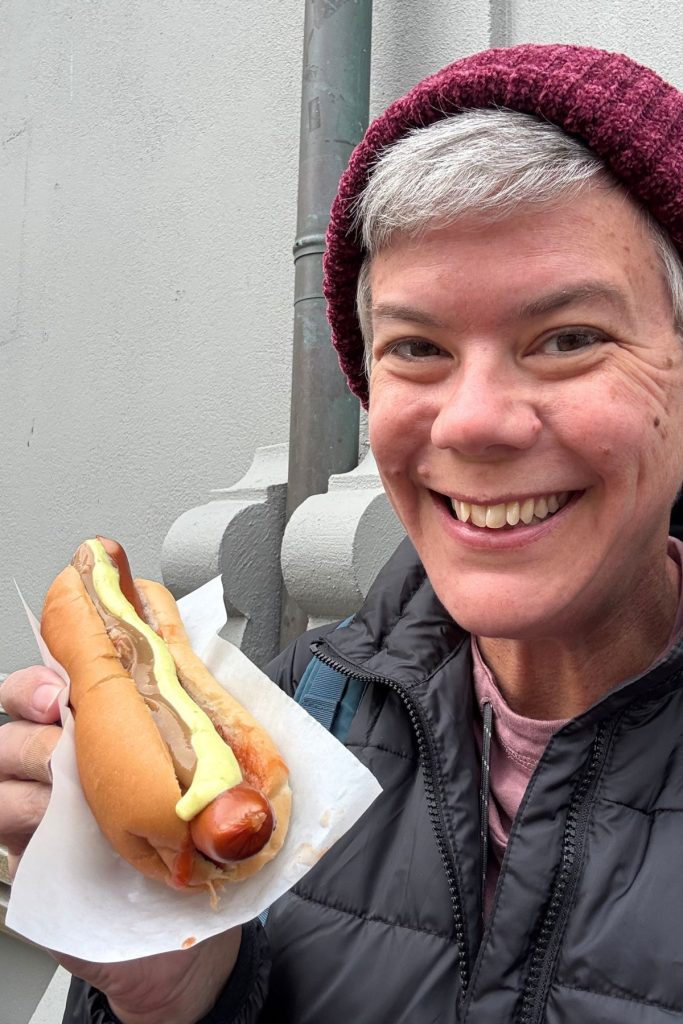 Smiling woman holding a hot dog with mustard and sauce in paper, in front of a gray wall