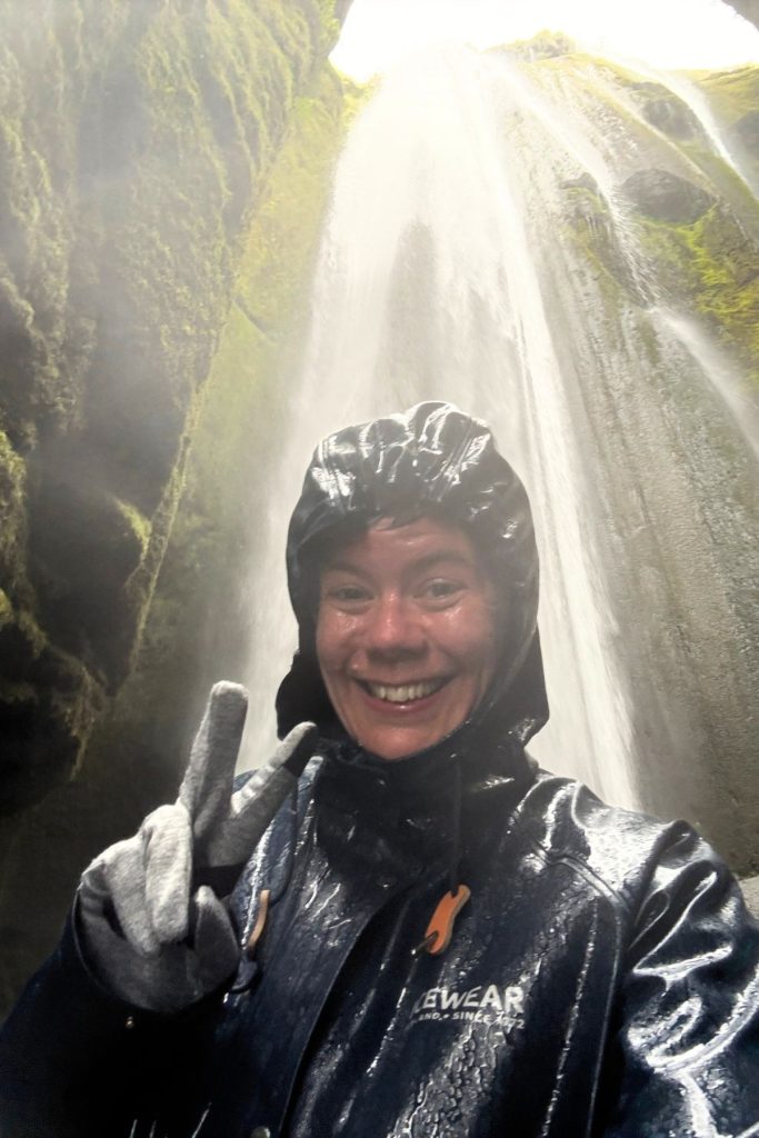 Woman in wet rain gear smiling in front of Gljúfrabúi waterfall in a moss-covered canyon