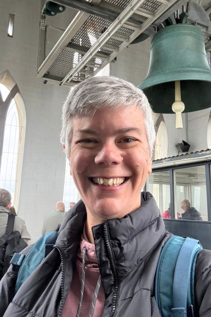Smiling woman in a gray puffer jacket and backpack beneath a large green bell in the Hallgrímskirkja tower
