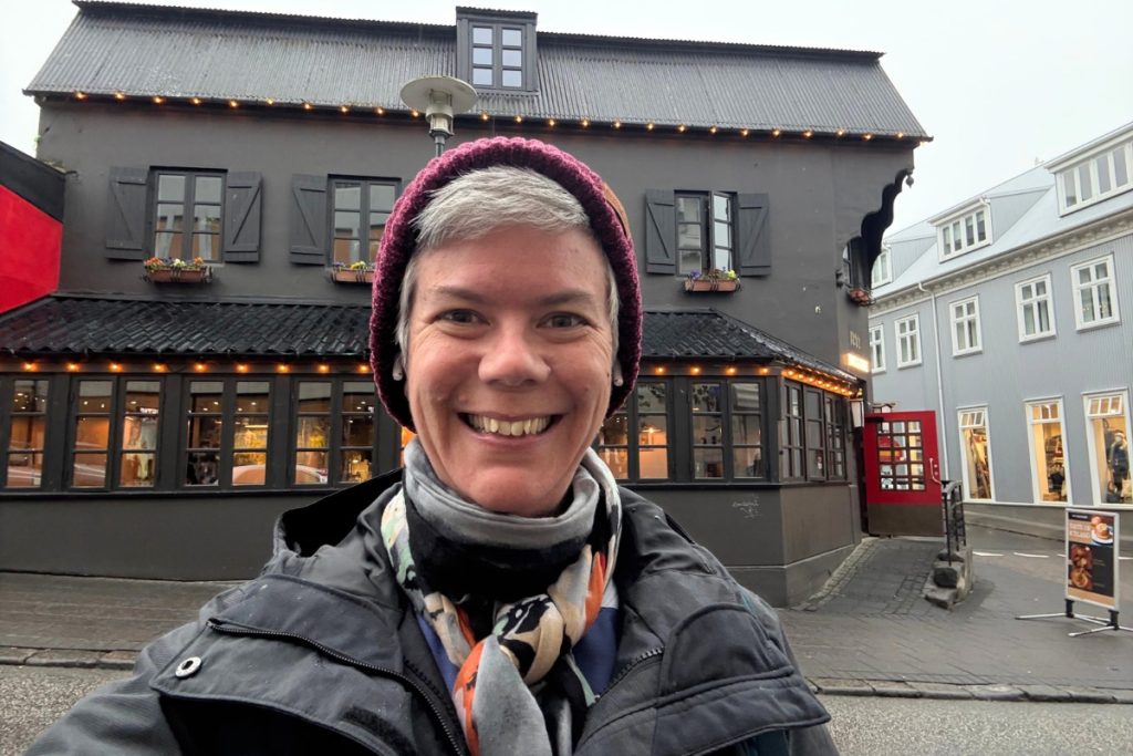 Smiling woman in a burgundy hat and scarf in front of a dark two-story restaurant with string lights
