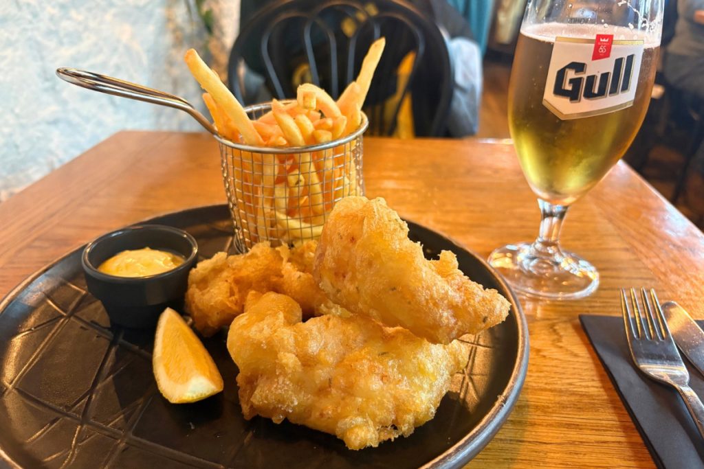 Battered fish pieces and fries in a wire basket with a dip and lemon wedge beside a glass of Gull beer