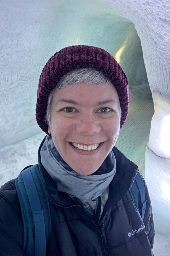 Smiling woman in a knit hat and scarf inside a pale blue-white ice cave at Perlan