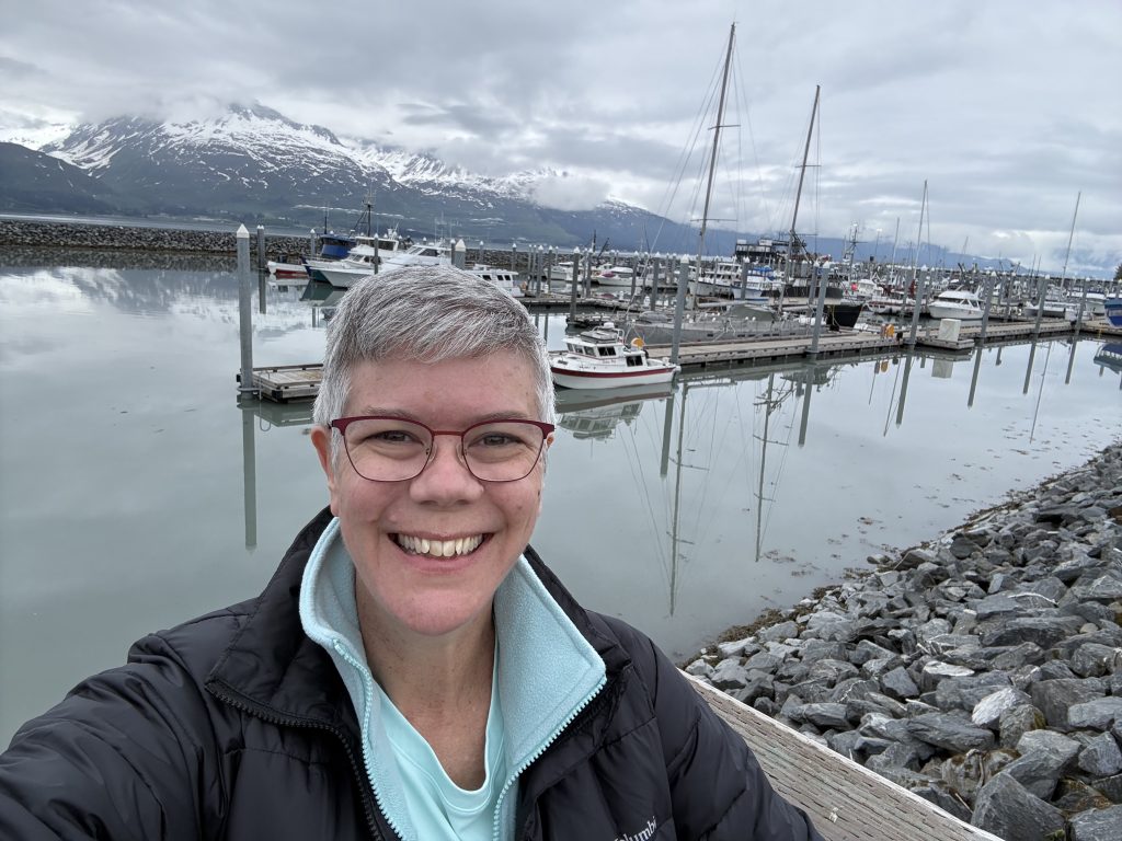 Selfie at Valdez harbor with fishing boats and snow-capped mountains in the background