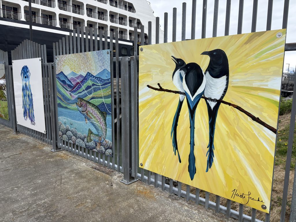 Paintings by local artists on display at the Valdez cruise terminal, featuring sea otters, salmon, and magpies