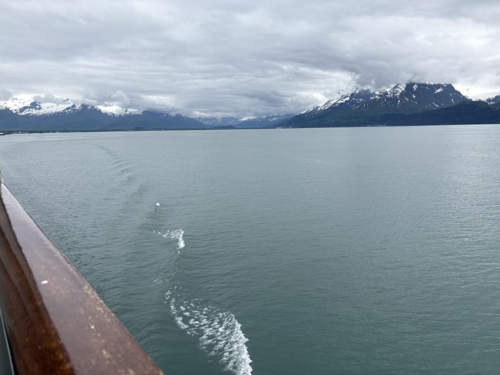Ship's wake trailing behind as the vessel sails out of Valdez, Alaska, with mountains in the distance