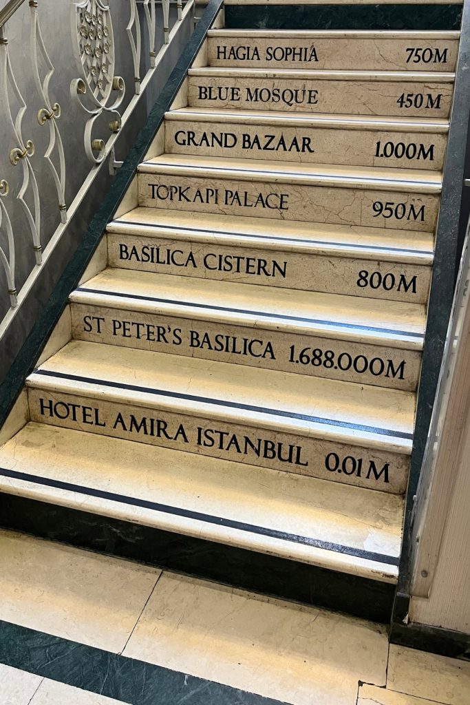 Marble hotel staircase with each step engraved listing distances to nearby landmarks including Hagia Sophia, Blue Mosque, Grand Bazaar, Topkapi Palace, Basilica Cistern, St. Peter's Basilica, and Hotel Amira Istanbul at 0.01 meters.