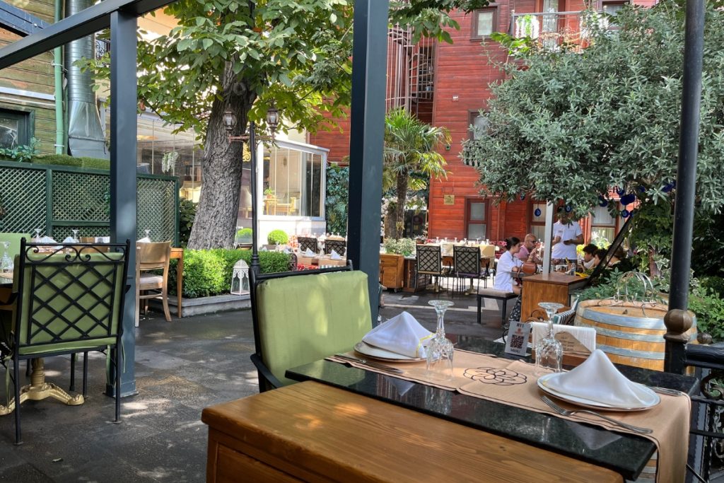 Outdoor restaurant courtyard with green metal chairs, wooden tables set with white napkins and glasses, string lights, and a large tree providing shade.