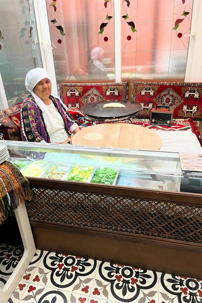 Woman in traditional dress preparing gözleme dough at a round griddle table, with a display case of fillings including cheese, spinach, and potato in front of her.
