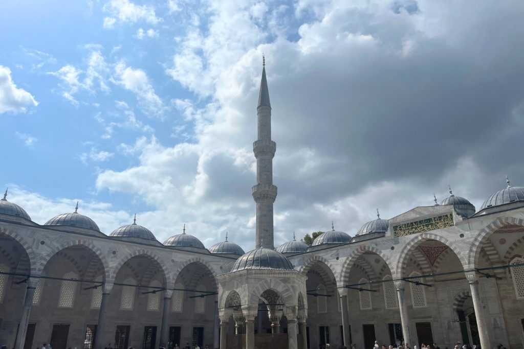 Courtyard of the Blue Mosque with a single tall minaret rising against a partly cloudy sky, domed arcades and people walking below.