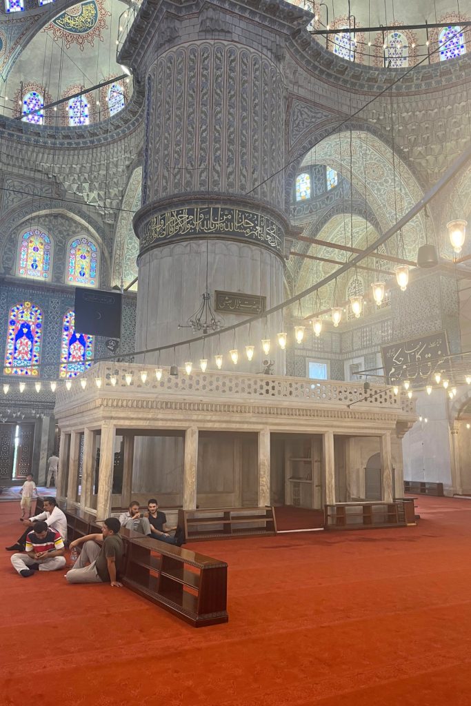 Interior of the Blue Mosque showing a raised marble platform with columns, surrounded by visitors sitting on a red carpeted floor beneath a large central dome.