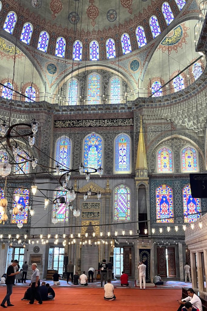 Wide interior view of the Blue Mosque showing stacked domes with stained glass windows, hanging chandeliers, and visitors sitting on a red carpeted floor.