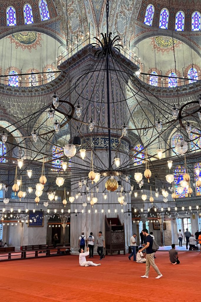 Interior view of the Blue Mosque with ornate gold chandeliers hanging low over a red carpeted floor, stained glass windows and a domed ceiling with Arabic calligraphy visible above.