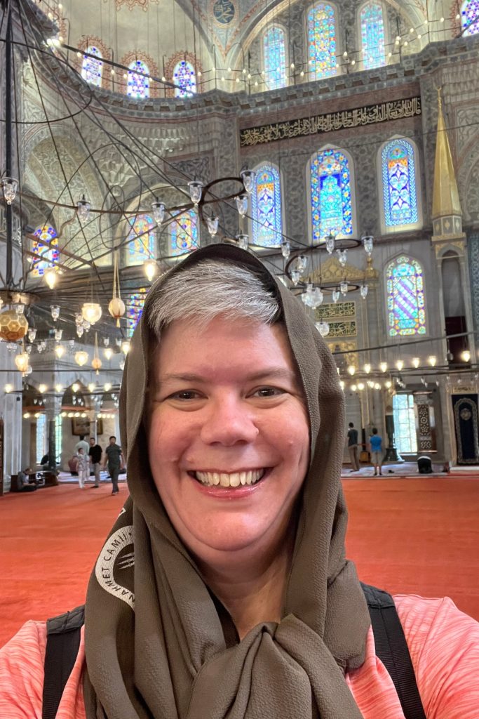 Selfie of a woman wearing a brown head covering smiling inside the Blue Mosque, with stained glass windows and hanging chandeliers visible behind her.