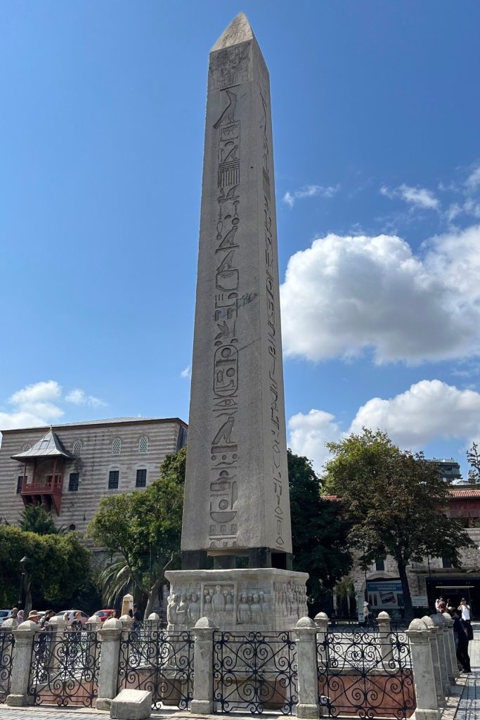 Tall carved stone Egyptian obelisk with hieroglyphic inscriptions, surrounded by an iron fence, with historic buildings and trees in the background.
