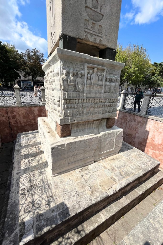 Detail of the carved marble pedestal beneath the Obelisk of Theodosius, showing relief sculptures of Byzantine figures and Latin inscription below.