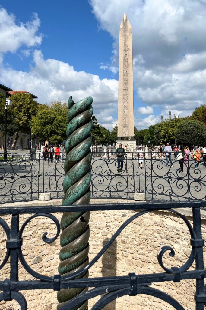 Twisted bronze Serpent Column in the foreground with the tall stone Obelisk of Theodosius visible in the background, framed by wrought iron fencing under a partly cloudy sky.