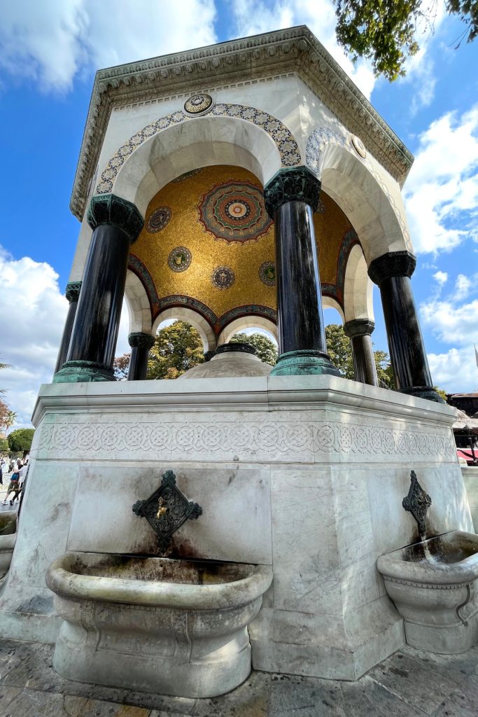 Ornate domed fountain pavilion with black marble columns and a gold mosaic ceiling, part of the German Fountain in Sultanahmet Square.